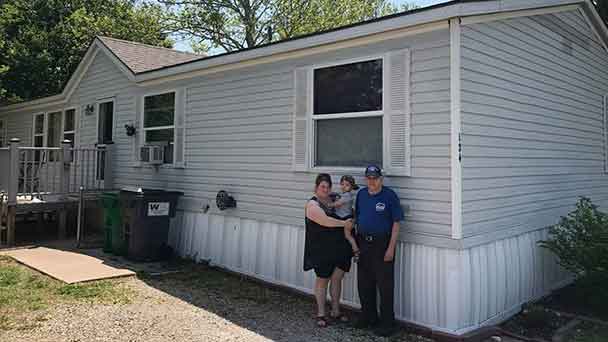 Three people stand together in front of a light gray manufactured home with white trim on a sunny day. Three people stand together in front of a light gray manufactured home with white trim on a sunny day.