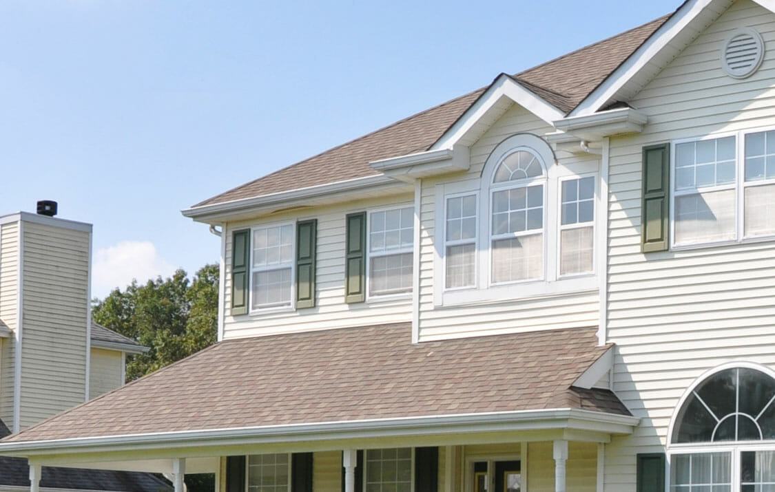 Two-story house with light-colored siding, green window shutters, and a covered porch under a clear blue sky.