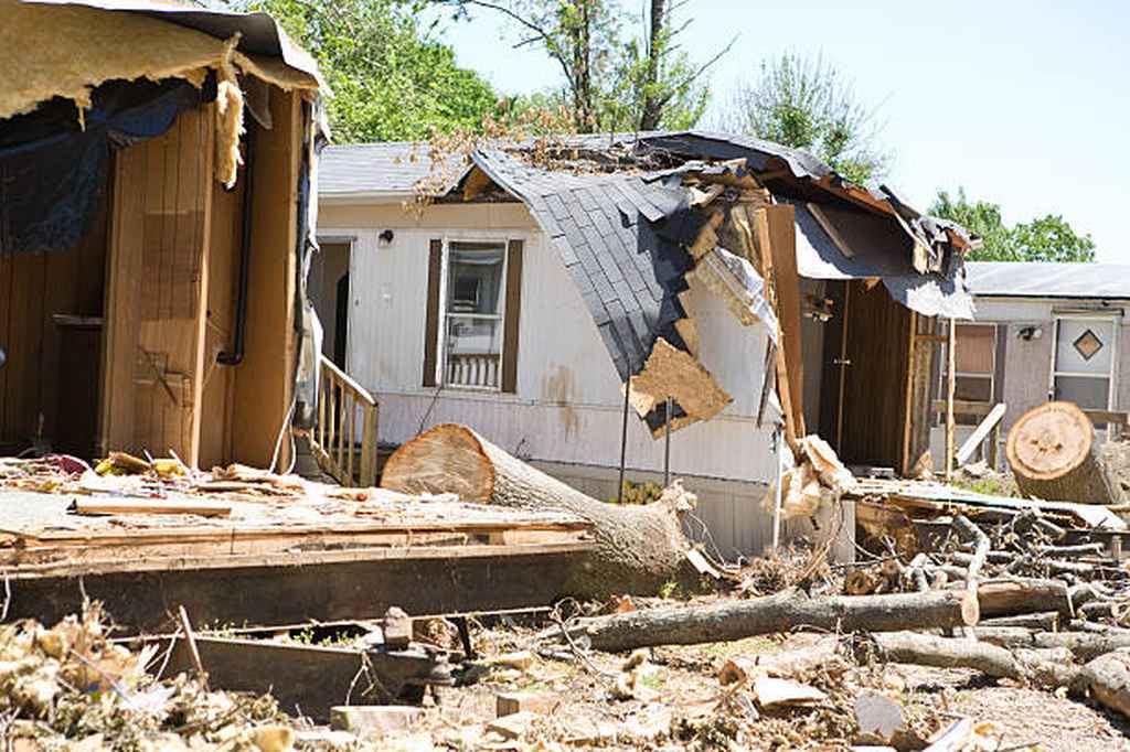 A mobile home with severe roof and wall damage, surrounded by fallen trees and debris, likely caused by a recent storm. A mobile home with severe roof and wall damage, surrounded by fallen trees and debris, likely caused by a recent storm.