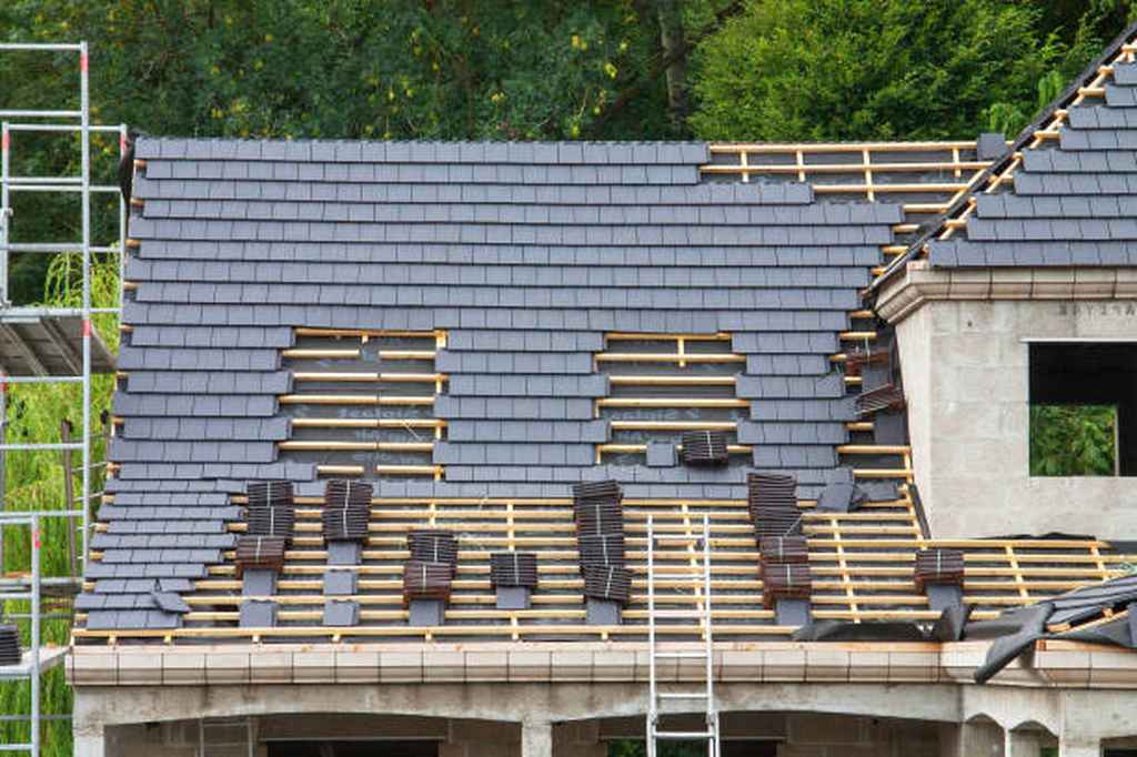A house roof under construction with dark roof tiles partially installed, wooden battens exposed, scaffolding and a ladder positioned nearby.