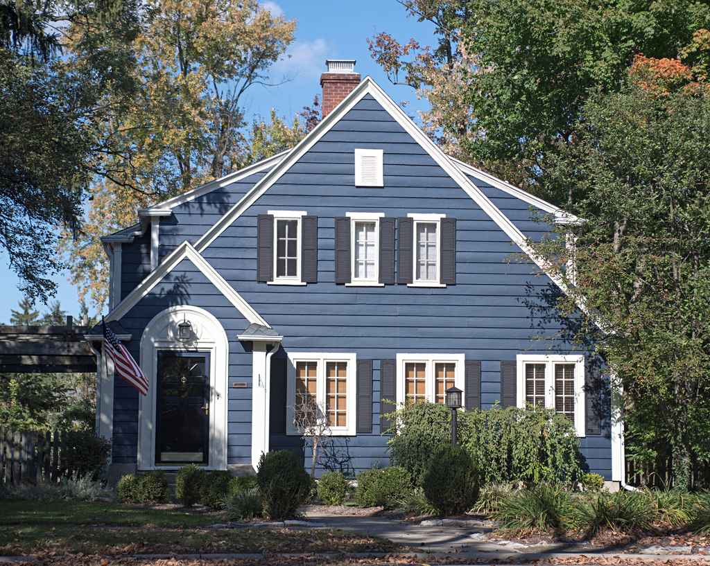 A blue two-story house with white trim, black shutters, and an American flag near the front door, surrounded by trees and shrubs on a sunny day. A blue two-story house with white trim, black shutters, and an American flag near the front door, surrounded by trees and shrubs on a sunny day.
