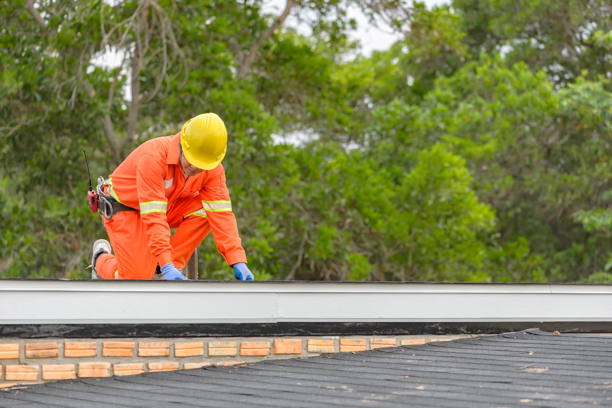 A worker in orange safety gear and a yellow helmet kneels on a roof, inspecting or repairing the edge, with trees visible in the background.
