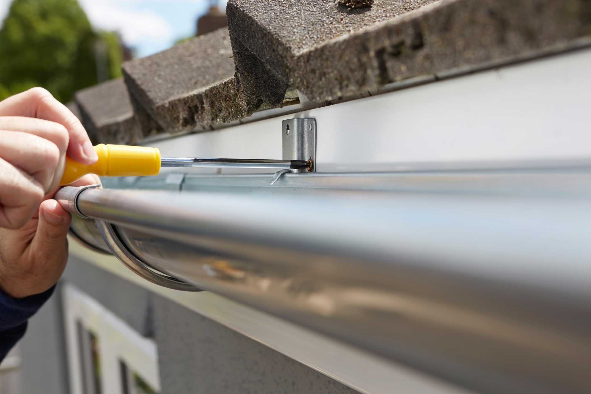 A person uses a screwdriver to attach a metal rain gutter to the side of a building under the edge of a roof.