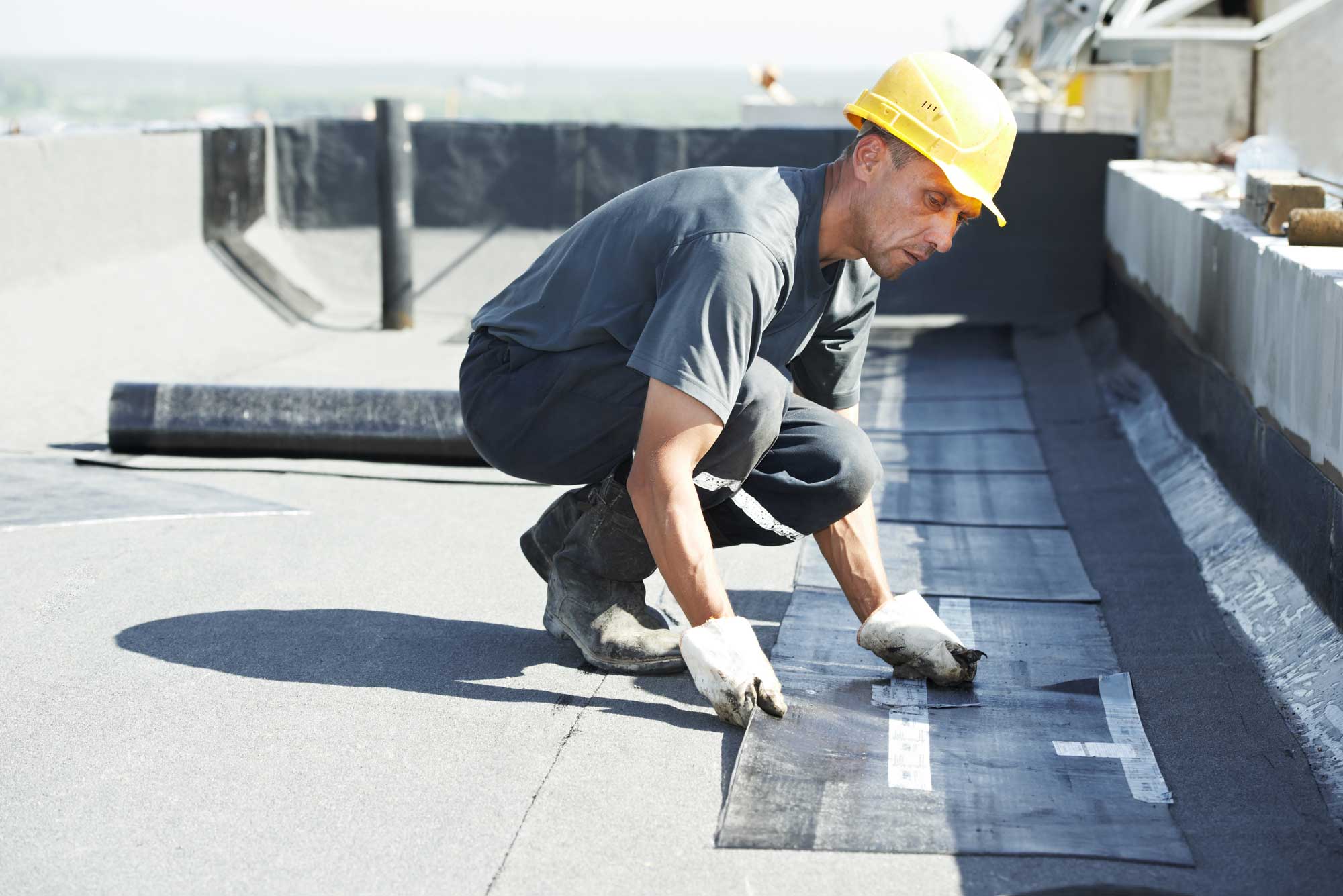 A construction worker wearing a yellow hard hat and gloves installs roofing material on a flat roof. A construction worker wearing a yellow hard hat and gloves installs roofing material on a flat roof.