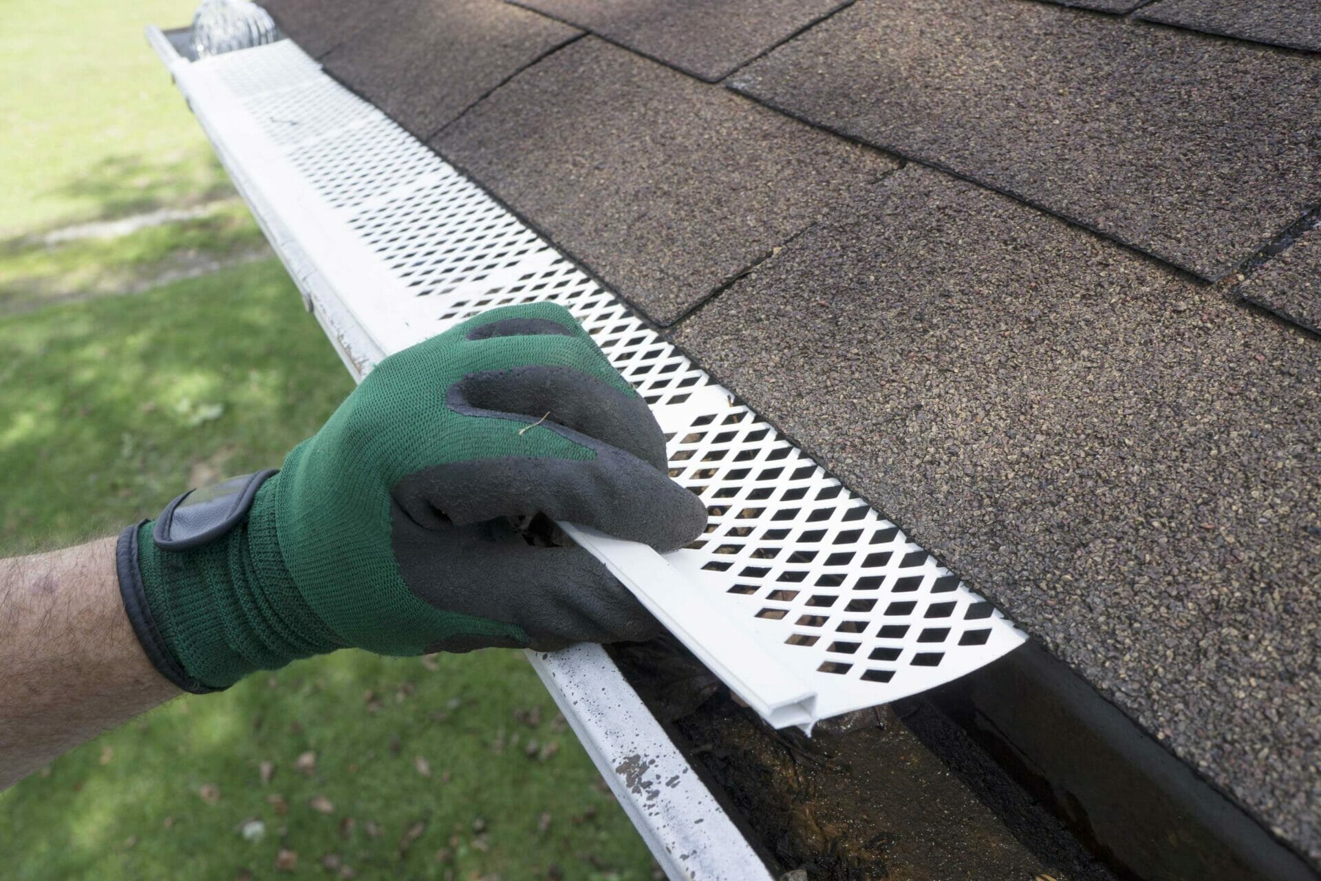 A person wearing a green glove is installing a white plastic gutter guard on the edge of a roof with brown shingles. A person wearing a green glove is installing a white plastic gutter guard on the edge of a roof with brown shingles.