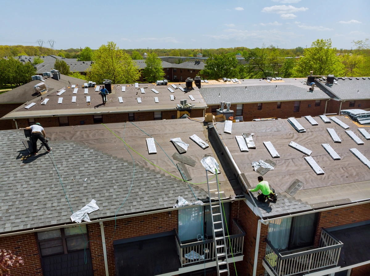 Workers are installing new roofing materials on apartment buildings; bundles of shingles and tools are spread across the rooftops under a clear sky.