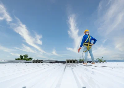 A construction worker wearing a safety harness and helmet stands on a white metal roof under a blue sky with scattered clouds.