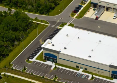 Aerial view of a large warehouse building with a white roof, multiple loading docks, parked trucks, and surrounding parking lots with landscaped greenery.