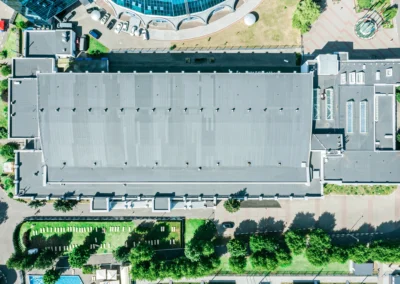 Aerial view of a large rectangular building with a gray roof, surrounded by trees, sidewalks, parked cars, and adjacent to a curved glass structure.