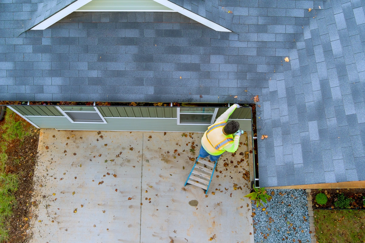 A person in a high-visibility vest stands on a ladder cleaning leaves from the gutter of a house with a gray shingle roof.