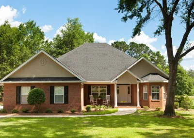 Single-story brick house with dark shutters, a covered front porch, rocking chairs, and a well-maintained lawn surrounded by trees on a sunny day.