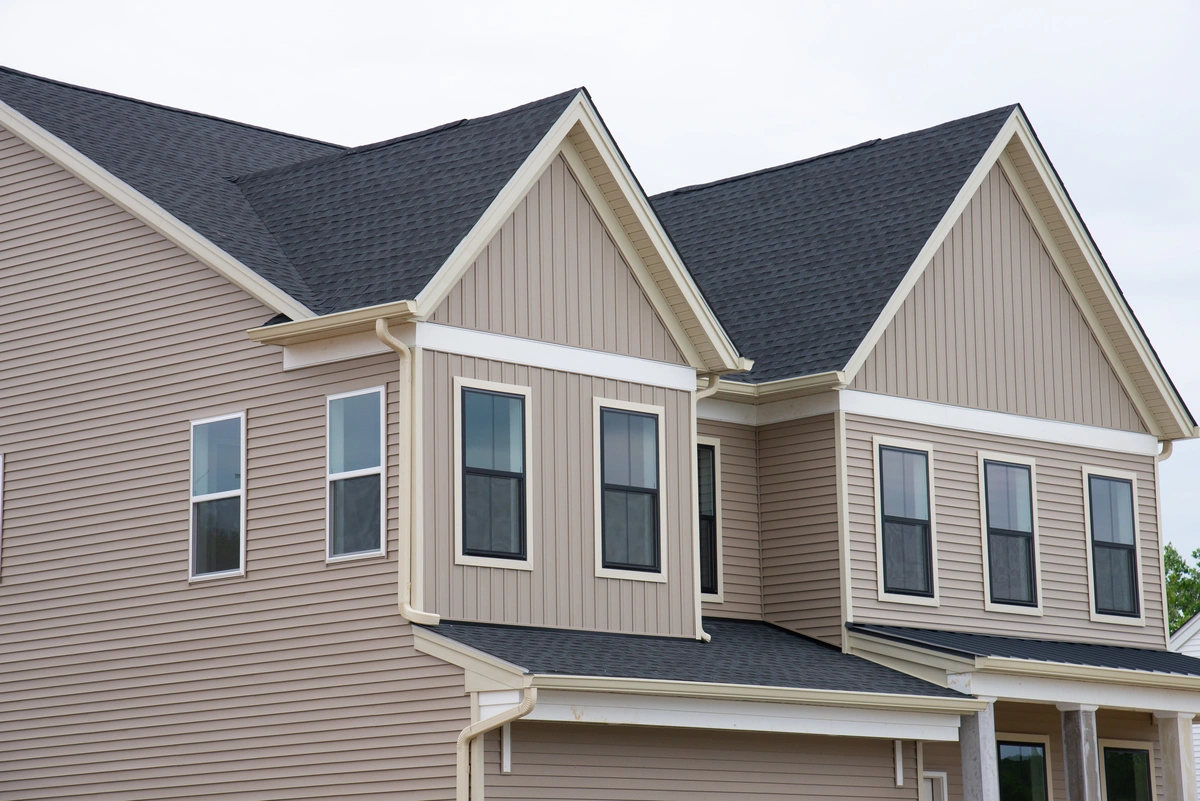 Two-story beige house with vinyl siding, multiple windows, white trim, and a dark shingled roof under a cloudy sky.