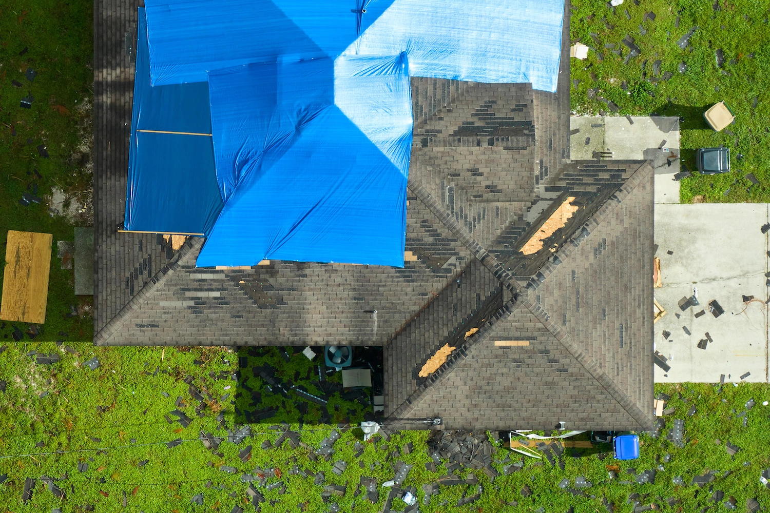 Aerial view of a house with damaged roof partially covered by blue tarps; shingles and debris scattered on the surrounding lawn and driveway.