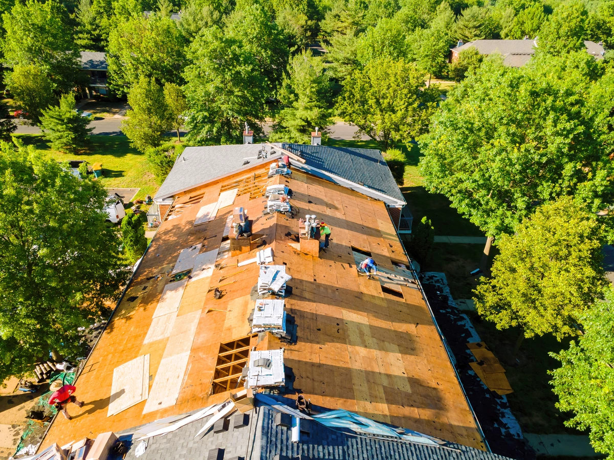 Aerial view of workers repairing or replacing shingles on a residential roof surrounded by trees in a suburban neighborhood. Aerial view of workers repairing or replacing shingles on a residential roof surrounded by trees in a suburban neighborhood.
