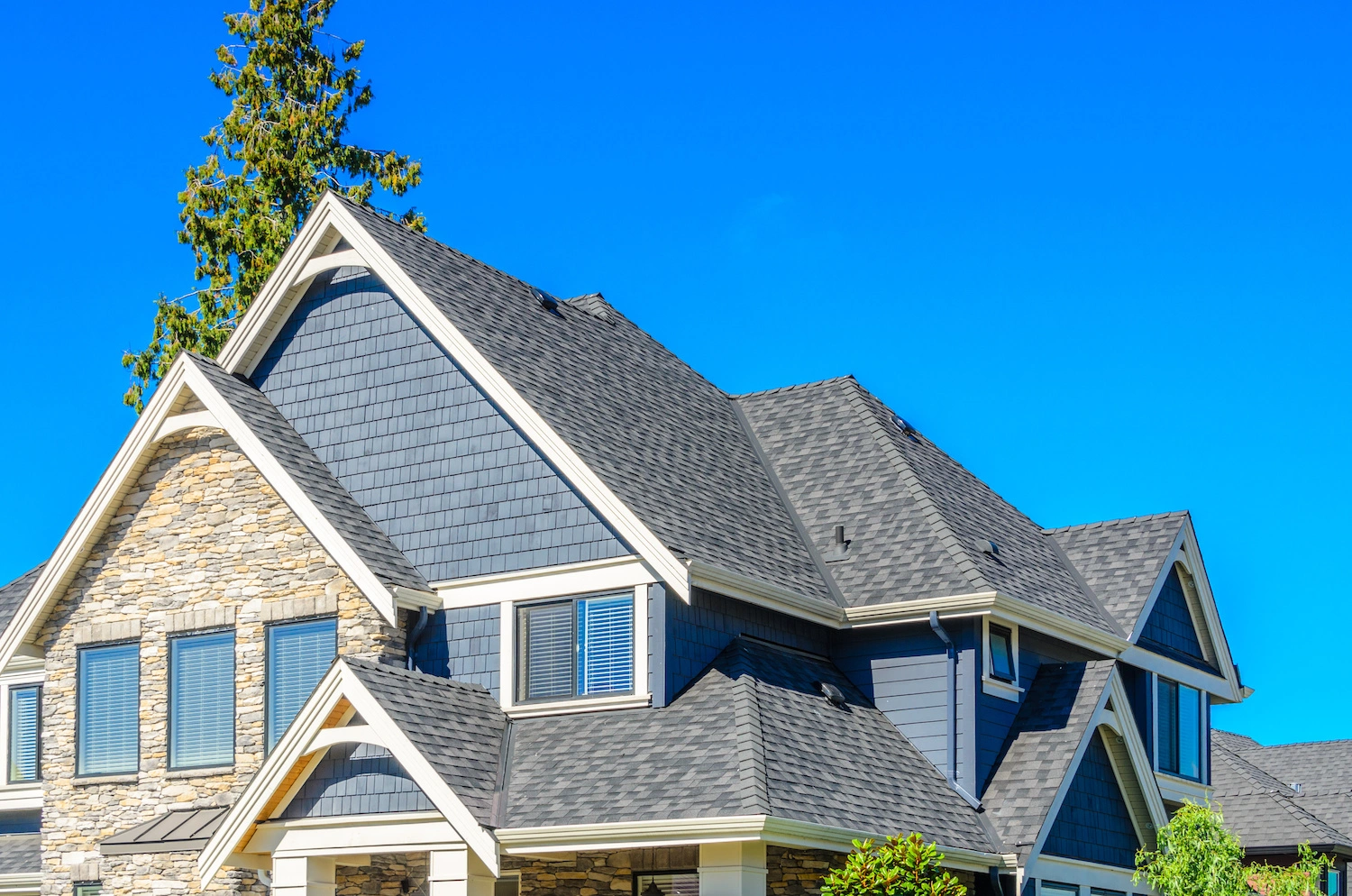 Modern two-story house with blue siding, stone accents, and dark gray shingle roof, set against a clear blue sky.