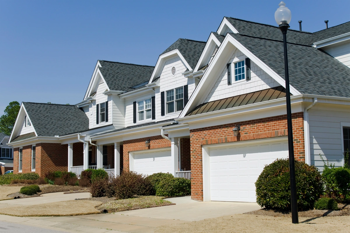 Row of two-story suburban townhouses with brick and white siding exteriors, each with a double garage, front shrubs, and a sidewalk under a clear sky.