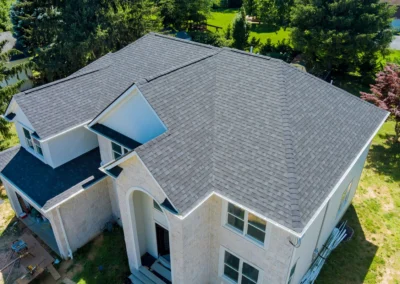 Aerial view of a two-story house with light-colored exterior walls and a dark gray shingle roof surrounded by green trees and lawn.