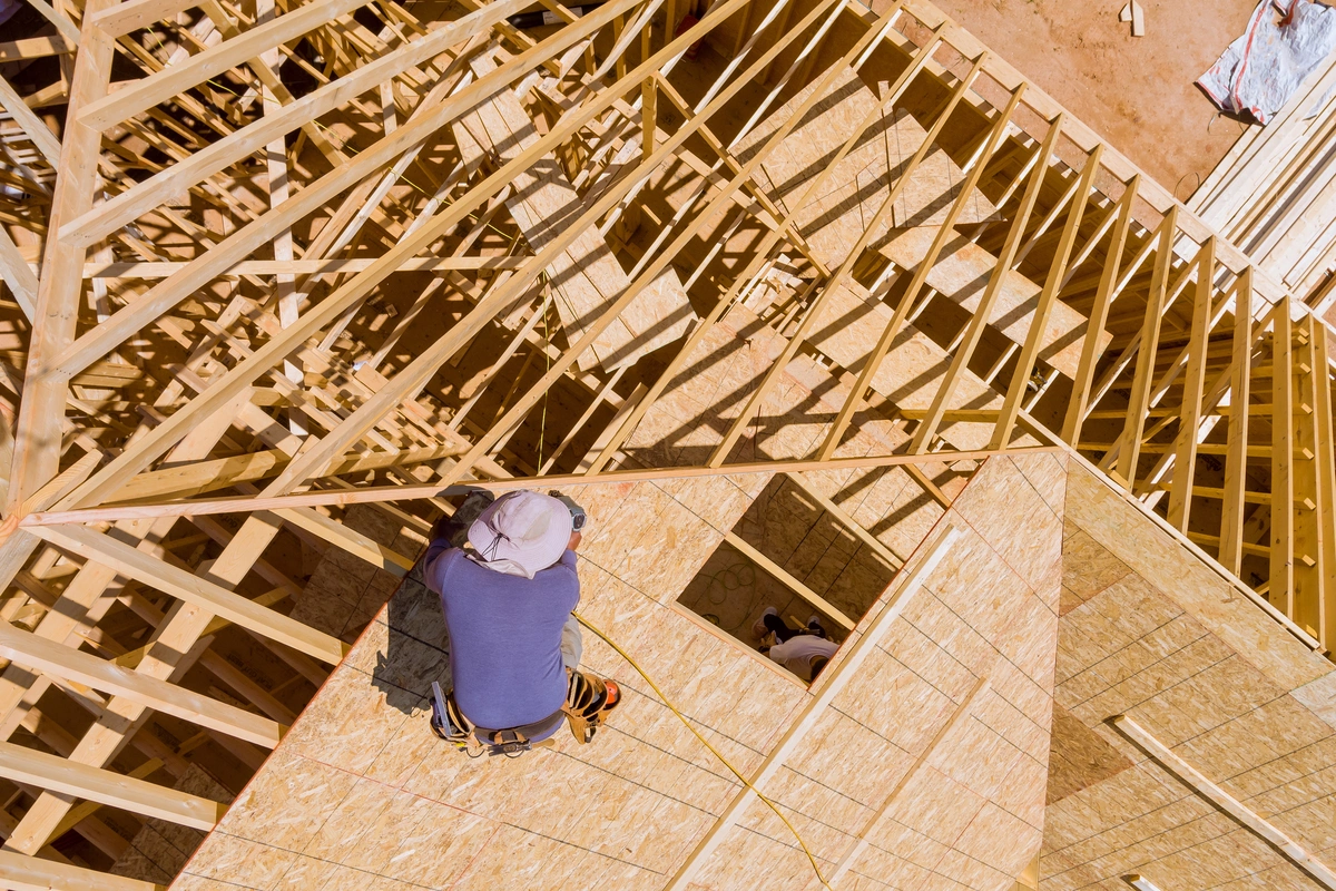 A construction worker wearing a hat is assembling the wooden frame of a house roof from an overhead view.