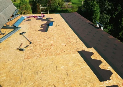 Plywood roof deck partially covered with new asphalt shingles, with tools and materials scattered on the surface under daylight.