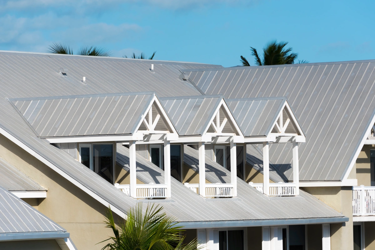 A row of dormer windows with white trim extends from a metal roof of a beige building, with palm trees and a blue sky in the background. A row of dormer windows with white trim extends from a metal roof of a beige building, with palm trees and a blue sky in the background.