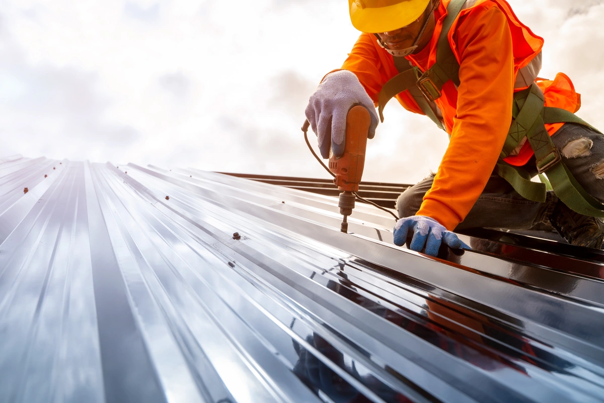 A construction worker in safety gear uses a power drill to fasten screws into a metal roof panel under a cloudy sky. A construction worker in safety gear uses a power drill to fasten screws into a metal roof panel under a cloudy sky.