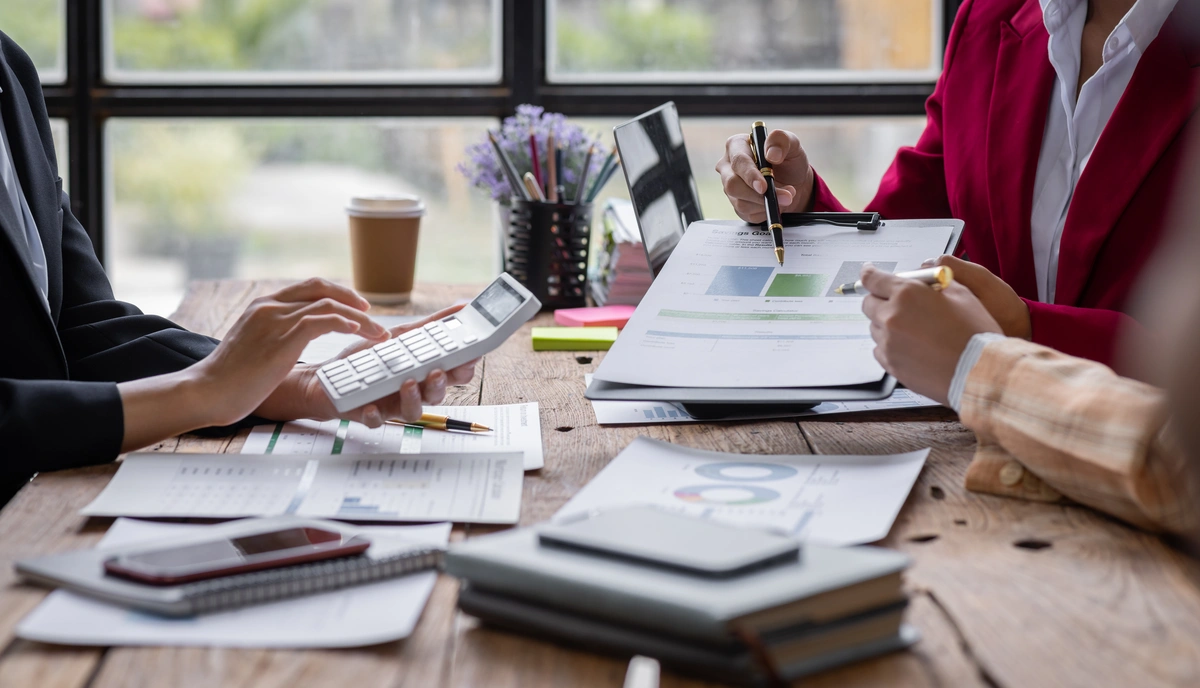 Three people sit at a table reviewing financial documents, using a calculator and writing on charts and graphs, with notebooks and coffee on the wooden surface. Three people sit at a table reviewing financial documents, using a calculator and writing on charts and graphs, with notebooks and coffee on the wooden surface.