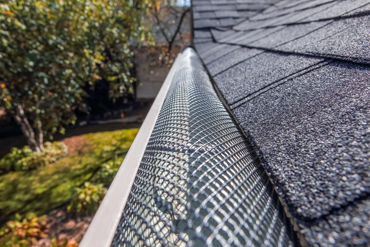 Close-up view of a mesh gutter guard installed along the edge of an asphalt shingle roof, with trees and a landscaped yard in the background. Close-up view of a mesh gutter guard installed along the edge of an asphalt shingle roof, with trees and a landscaped yard in the background.