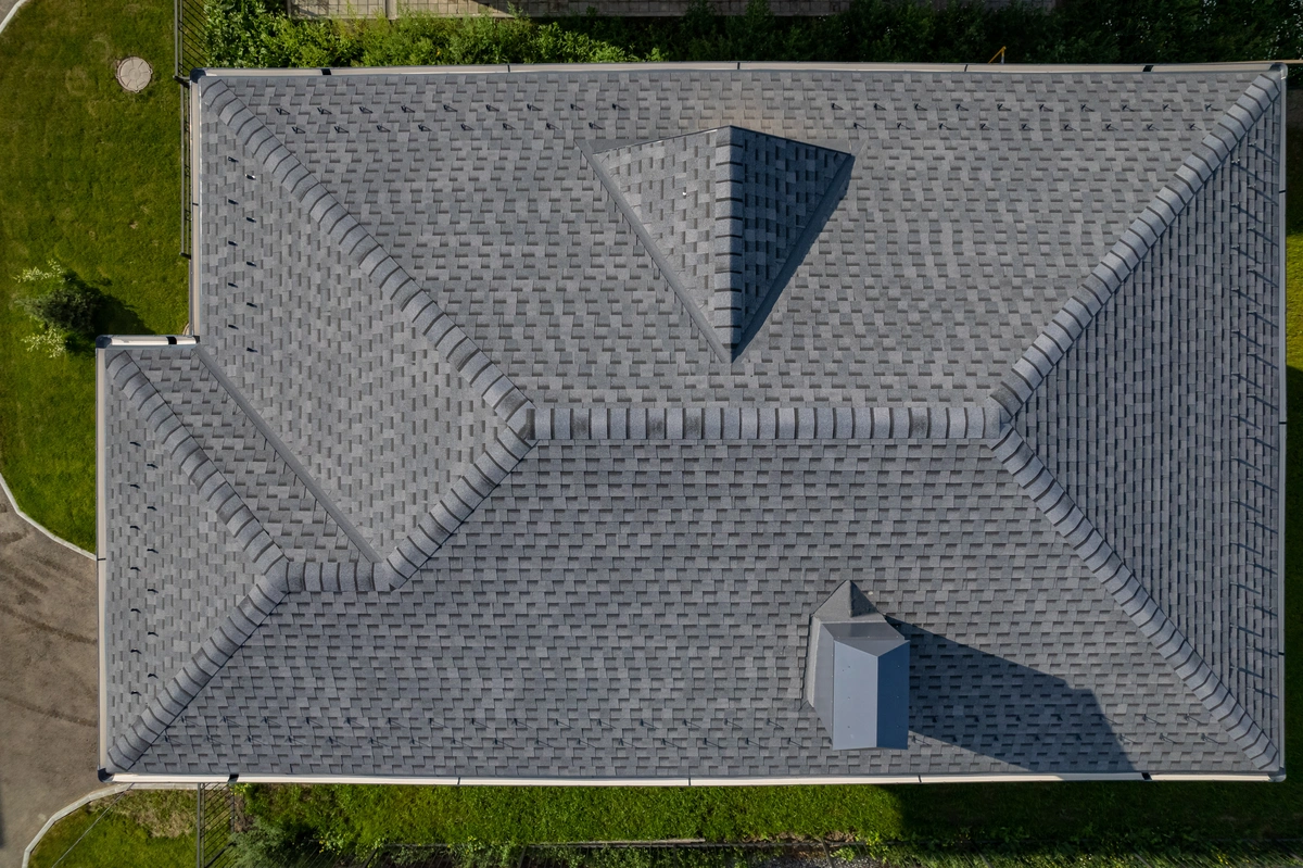 Aerial view of a gray shingled residential roof with multiple gables, hip features, and a small rooftop structure. Aerial view of a gray shingled residential roof with multiple gables, hip features, and a small rooftop structure.