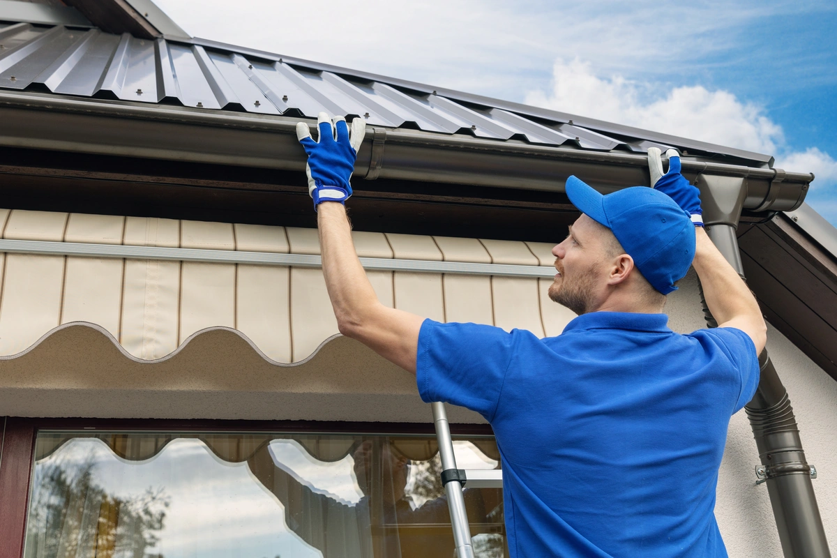 A man in blue work clothes and gloves stands on a ladder, installing or repairing a rain gutter on the roof edge of a house. A man in blue work clothes and gloves stands on a ladder, installing or repairing a rain gutter on the roof edge of a house.
