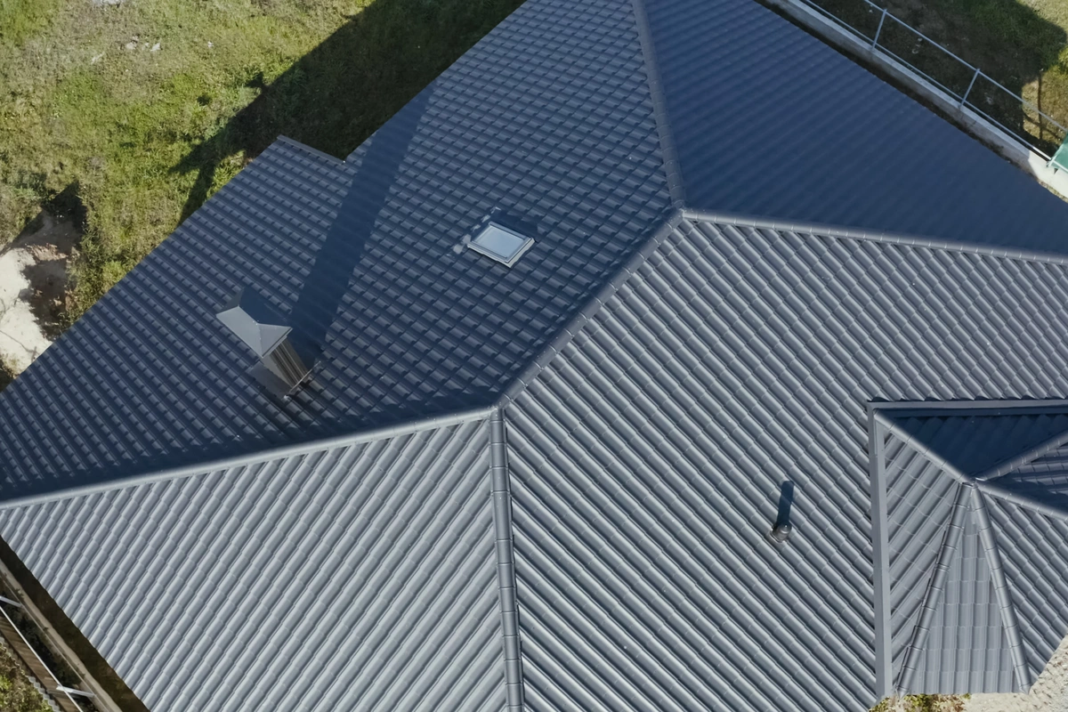 Aerial view of a building with a dark gray, metal, hipped roof featuring one skylight and two vents, surrounded by grass and a partially visible fence. Aerial view of a building with a dark gray, metal, hipped roof featuring one skylight and two vents, surrounded by grass and a partially visible fence.