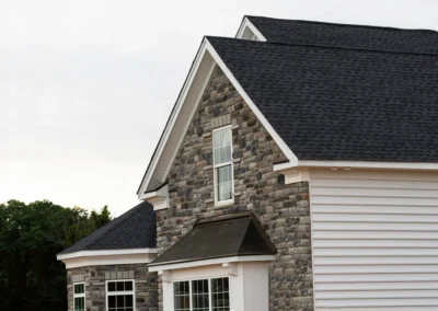 A two-story house with stone and white siding exterior, multiple windows, and a dark shingled roof against a backdrop of trees.