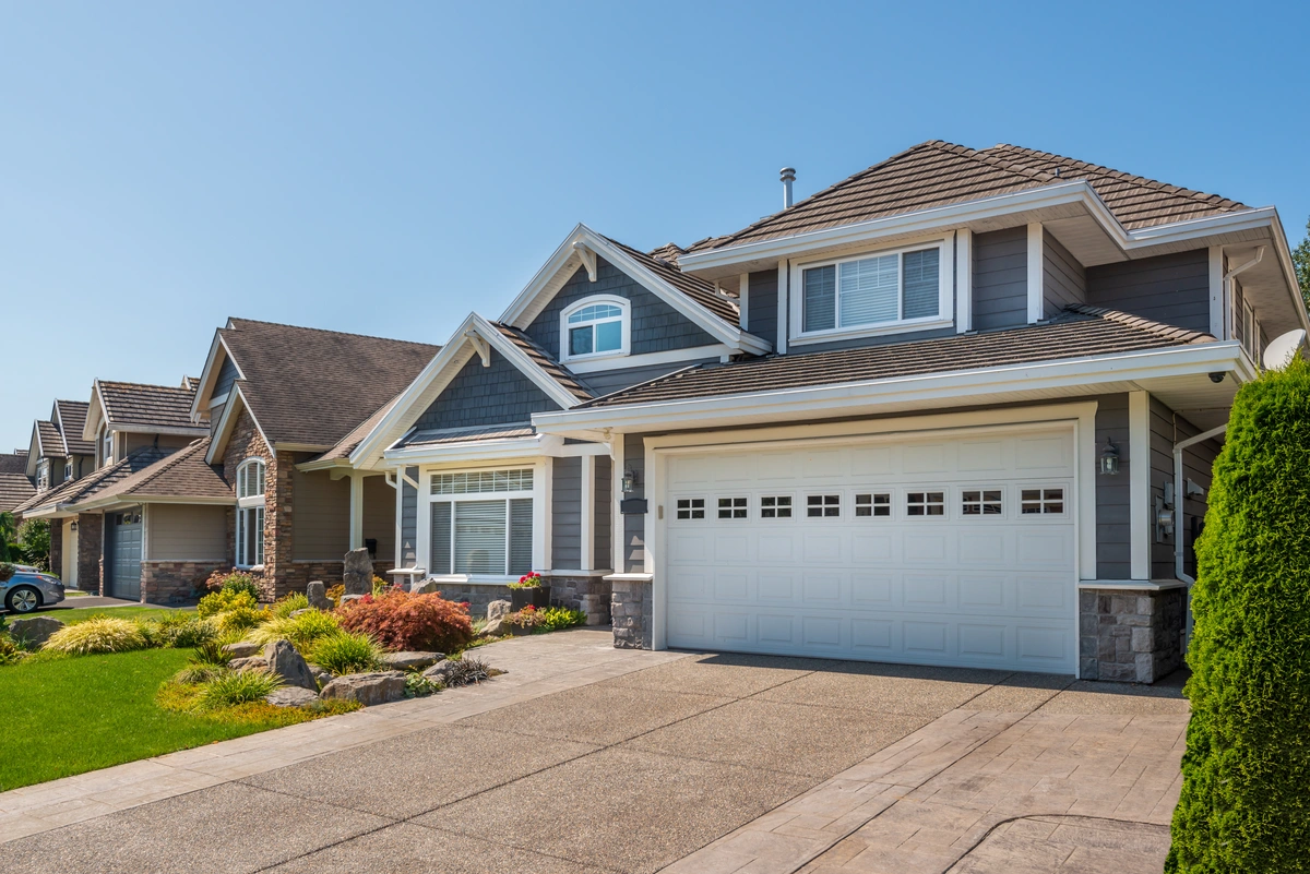 Suburban house with gray siding, white trim, a double garage door, manicured lawn, and landscaping, photographed on a clear, sunny day. Suburban house with gray siding, white trim, a double garage door, manicured lawn, and landscaping, photographed on a clear, sunny day.