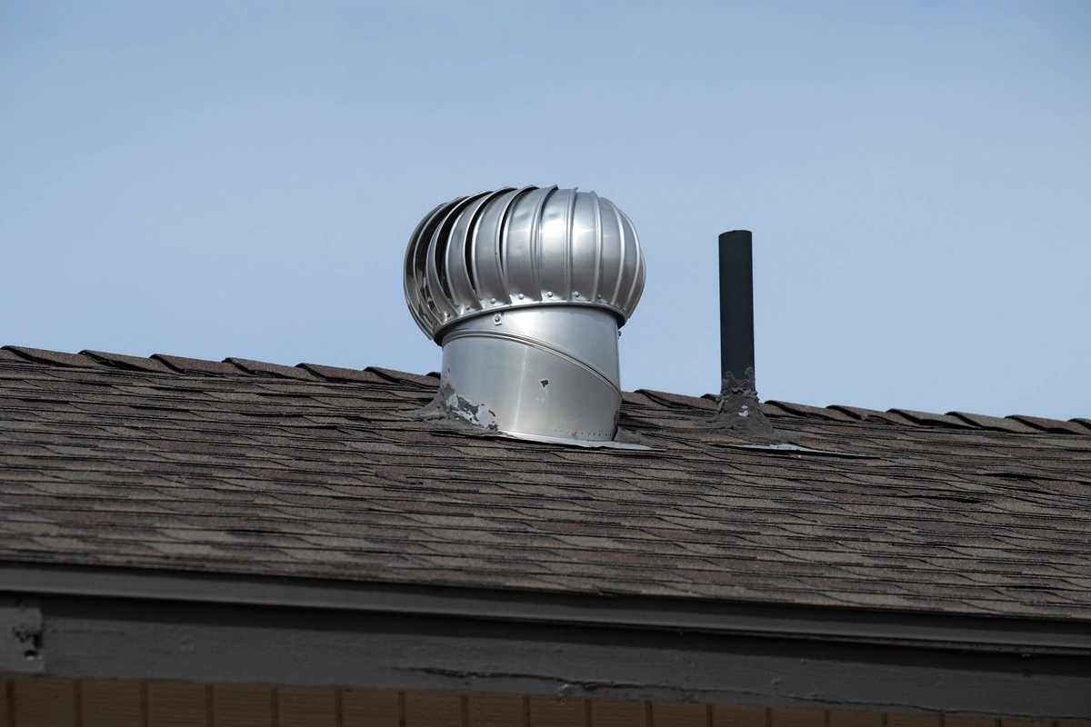A metal turbine roof vent and a black pipe are installed on a shingled roof under a clear blue sky. A metal turbine roof vent and a black pipe are installed on a shingled roof under a clear blue sky.