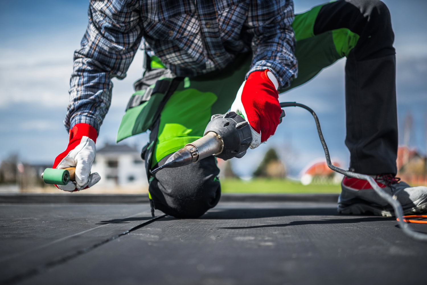 Worker in protective gloves kneeling on a flat roof, using a heat welding tool to seal roofing membrane seams. Worker in protective gloves kneeling on a flat roof, using a heat welding tool to seal roofing membrane seams.