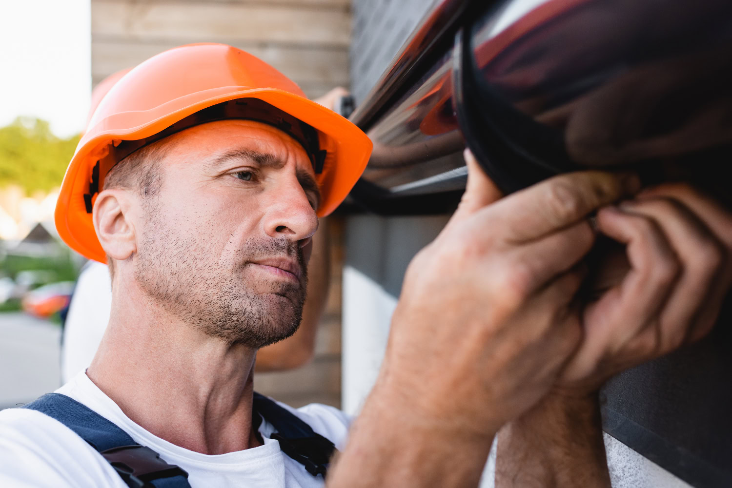 Man wearing an orange hard hat and white shirt installs or inspects a black fixture on an exterior wall, focusing intently on his work. Man wearing an orange hard hat and white shirt installs or inspects a black fixture on an exterior wall, focusing intently on his work.