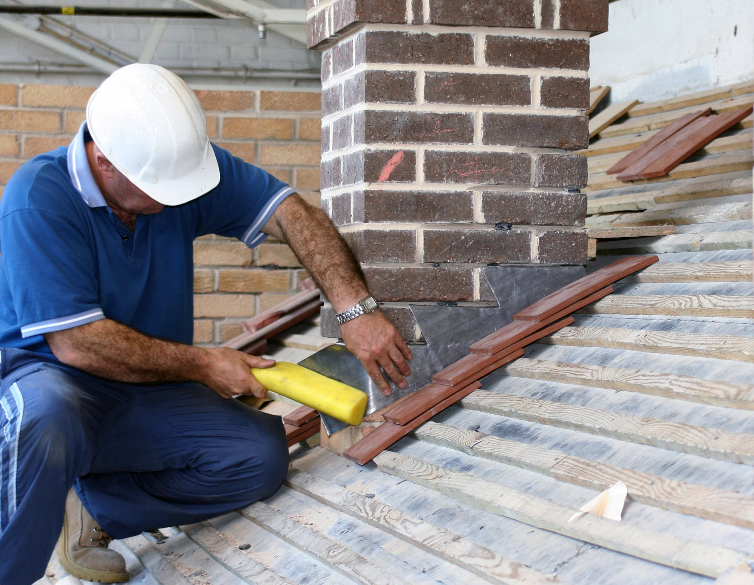 A construction worker in a white hard hat installs roof tiles around a brick chimney on a sloped wooden roof. A construction worker in a white hard hat installs roof tiles around a brick chimney on a sloped wooden roof.