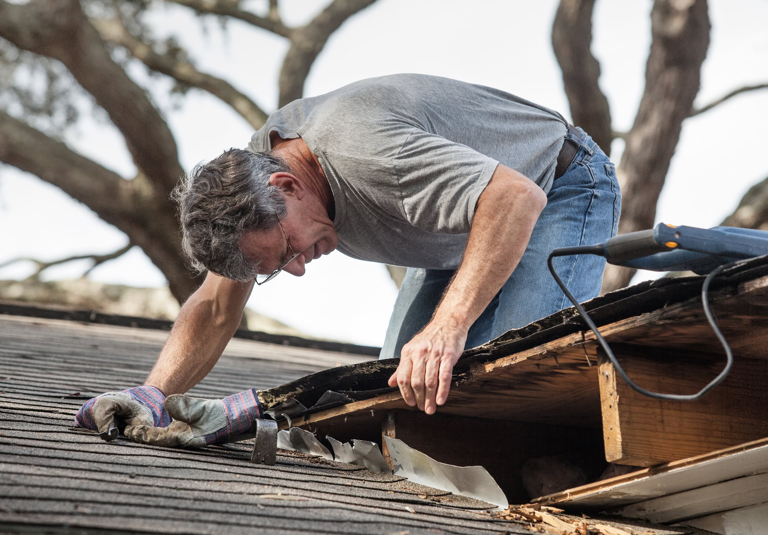 A man in work gloves inspects and repairs damaged shingles and wood on the edge of a roof, with a power tool nearby. A man in work gloves inspects and repairs damaged shingles and wood on the edge of a roof, with a power tool nearby.