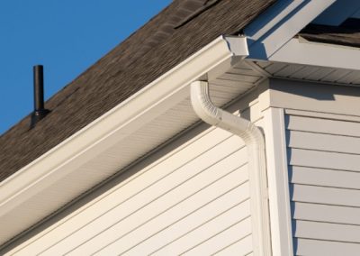 Close-up of a house corner showing grey asphalt shingles, white siding, a rain gutter, and a downspout against a clear blue sky.