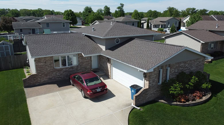 A single-story suburban house with brick and siding exterior, an attached two-car garage, and a red car parked in the driveway. Green lawns and neighboring houses are visible.