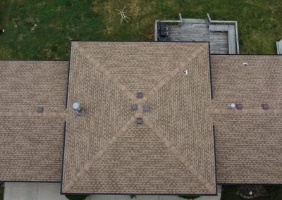 Aerial view of a house with a brown shingle roof, surrounded by grass, a wooden deck, and a small patio area in the backyard.