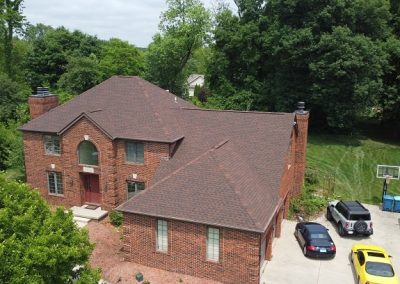 A two-story red brick house with a brown roof, surrounded by trees, with three cars and a basketball hoop in the driveway.