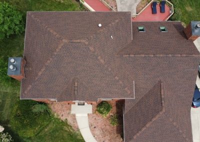 Aerial view of a house with a brown shingled roof, surrounding lawn, a curved walkway, deck with patio furniture, and several cars parked in the driveway.
