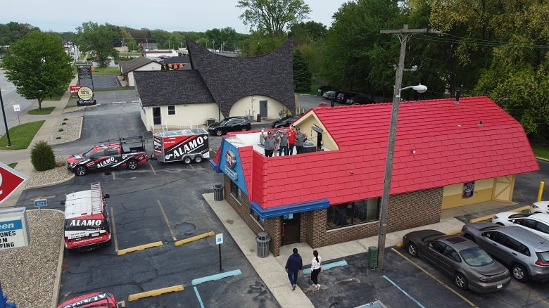 Aerial view of a restaurant with a red roof and parking lot; several Alamo-branded vehicles are parked outside and a group of people stands on the roof.