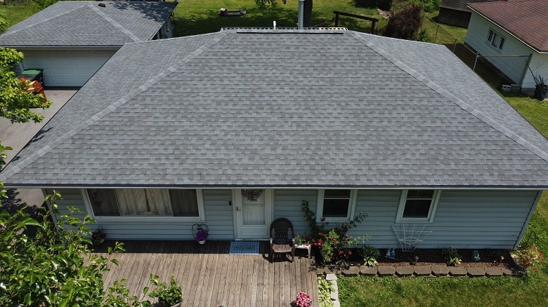 Aerial view of a single-story house with a gray shingled hip roof, light siding, front porch, garden beds, and a detached garage in the background.