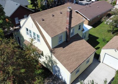 Aerial view of a beige two-story house with a brown roof, brick chimney, detached garage, and surrounding trees in a residential neighborhood.