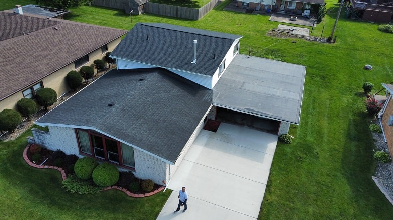 Aerial view of a single-story house with a dark roof, attached carport, concrete driveway, and maintained lawn; a person stands near the entrance. Aerial view of a single-story house with a dark roof, attached carport, concrete driveway, and maintained lawn; a person stands near the entrance.