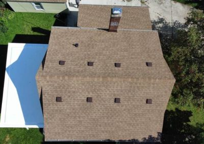 Aerial view of a house with a brown shingle roof, chimney, multiple roof vents, and a white flat-roofed extension on the left side.